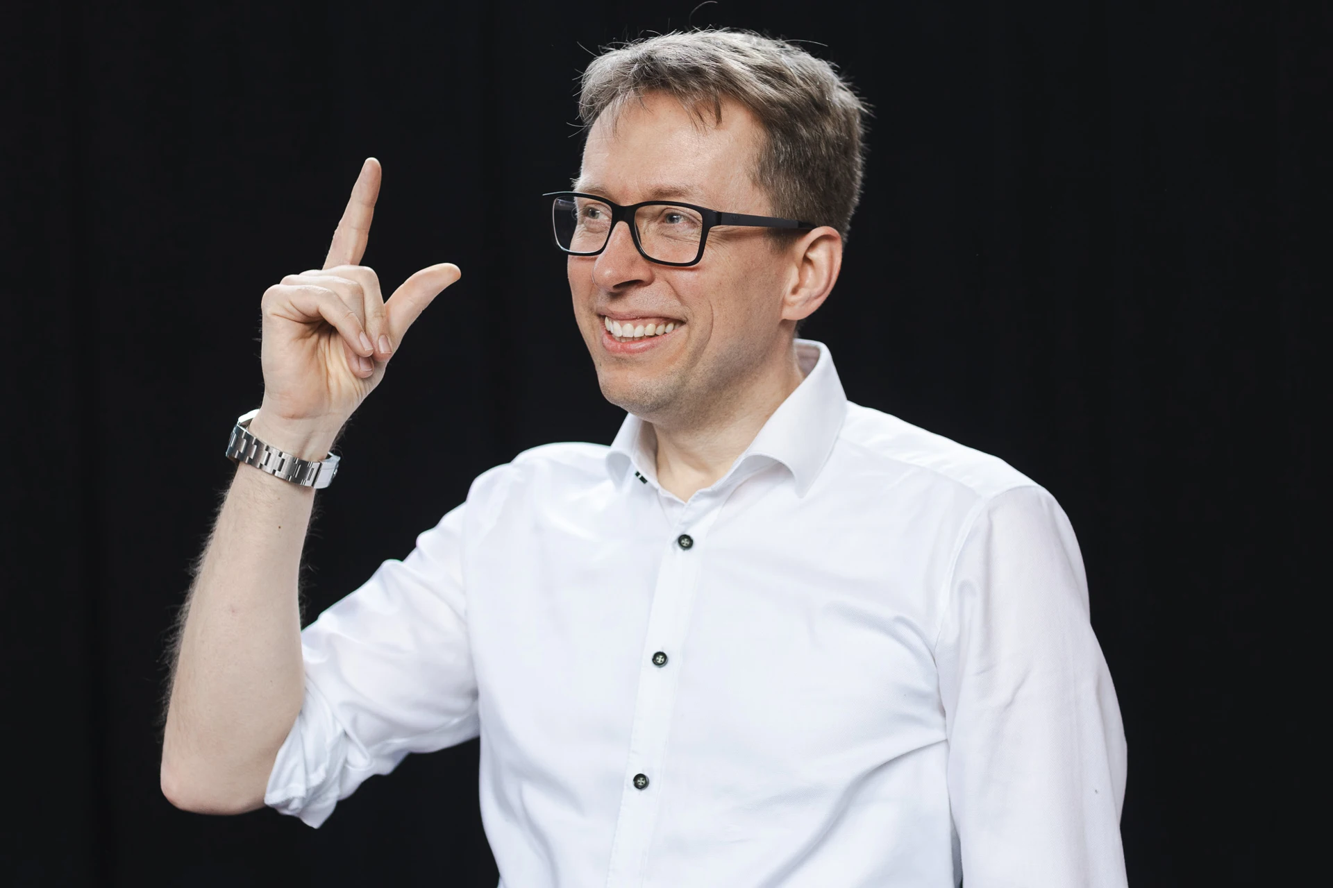 Daniel Schwenger lächelt und hebt einen Finger; er trägt ein weißes Hemd vor einem dunklen Hintergrund.